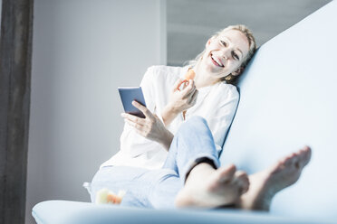 Laughing woman sitting on couch with cell phone eating a carrot - UUF11434