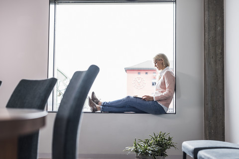 Geschäftsfrau sitzt auf der Fensterbank und benutzt ein Tablet, lizenzfreies Stockfoto
