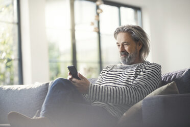 Senior man sitting on couch, using smartphone - SBOF00461