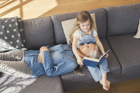 Grandmother and granddaughter sitting on couch, reading together a book stock photo