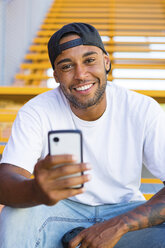 Porträt eines lächelnden jungen Mannes mit Smartphone auf einer Treppe sitzend - MGIF00059
