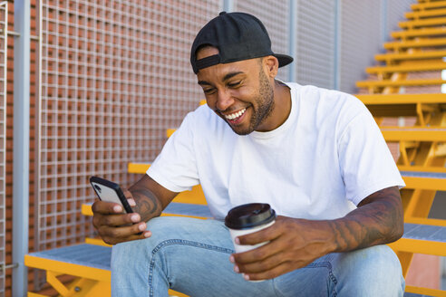 Lachender junger Mann mit Kaffee zum Mitnehmen, der auf einer Treppe sitzt und auf sein Smartphone schaut - MGIF00060