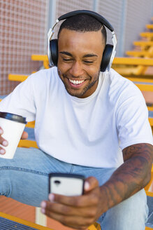Lachender junger Mann mit Kopfhörern und Kaffee zum Mitnehmen sitzt auf der Treppe und schaut auf sein Smartphone - MGIF00061
