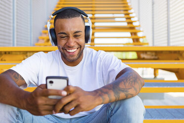 Porträt eines lachenden jungen Mannes mit Kopfhörern, der auf einer Treppe sitzt und auf sein Smartphone schaut - MGIF00063