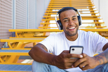 Portrait of laughing young man with headphones and smartphone sitting on stairs - MGIF00064