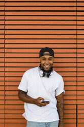 Porträt eines lachenden jungen Mannes mit Kopfhörern und Smartphone - MGIF00065