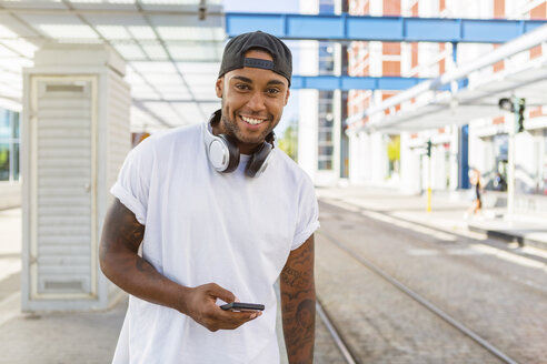 Porträt eines lächelnden jungen Mannes mit Kopfhörern und Smartphone, der an einer Straßenbahnhaltestelle wartet - MGIF00066