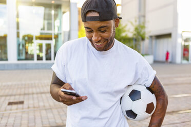 Lachender junger Mann mit Fußball und Blick auf sein Handy - MGIF00085