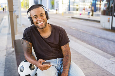 Portrait of smiling young man with soccer ball, headphones and cell phone waiting at tram stop - MGIF00094