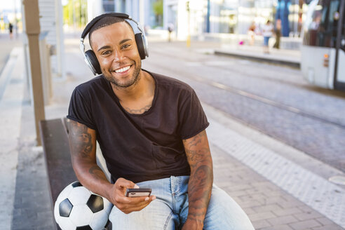 Porträt eines lächelnden jungen Mannes mit Fußball, Kopfhörern und Mobiltelefon, der an einer Straßenbahnhaltestelle wartet - MGIF00094
