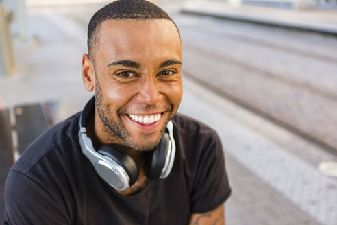 Porträt eines lachenden jungen Mannes mit Kopfhörern, der an einer Straßenbahnhaltestelle wartet - MGIF00095