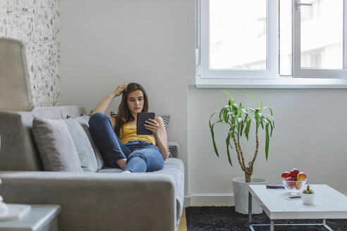 Frau auf Couch im Wohnzimmer beim Lesen eines E-Books - MOMF00211