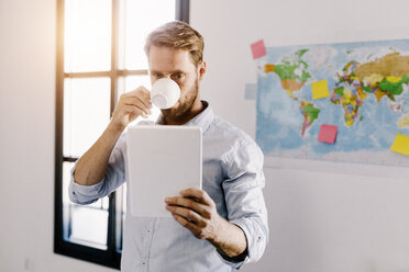 Bärtiger Mann im Büro trinkt Kaffee und schaut auf ein Tablet - GIOF03040