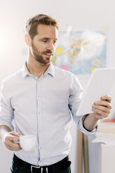 Bearded man in office with cup of coffee looking at tablet - GIOF03041
