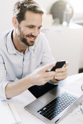 Porträt eines lächelnden Geschäftsmannes, der ein Smartphone am Schreibtisch in seinem Büro benutzt - GIOF03059