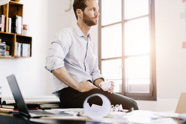 Architect with cup of coffee sitting on desk in his office looking at distance - GIOF03061