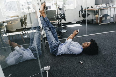 Junge Frau liegt auf dem Boden im Büro und überprüft ihr Handy, lizenzfreies Stockfoto
