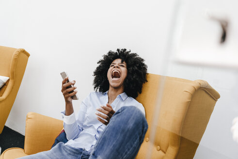 Screaming young woman with cell phone sitting in armchair - KNSF02282