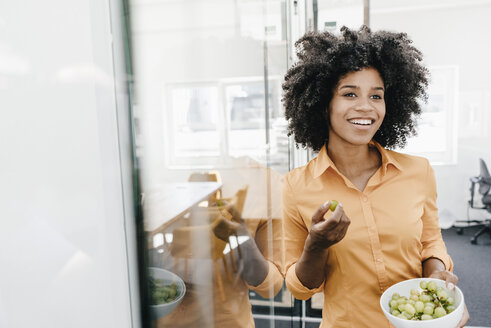 Smiling young woman holding bowl with grapes in office - KNSF02289