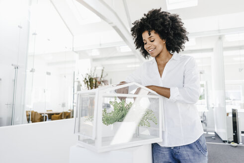 Smiling young woman in office caring for plants in glass box - KNSF02325