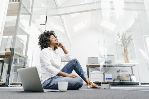 Junge Frau mit geschlossenen Augen sitzt auf dem Boden im Büro mit Laptop, lizenzfreies Stockfoto