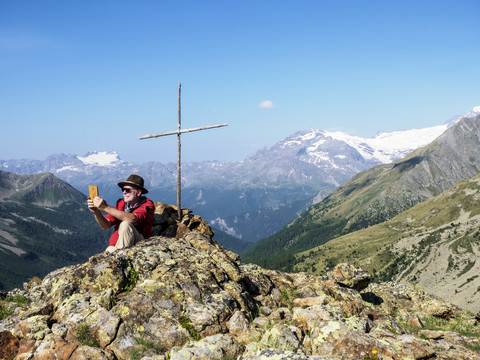 Italien Lombardei, Passo di Val Viola, Mann fotografiert mit seinem Smartphone, sitzend am Gipfelkreuz, lizenzfreies Stockfoto