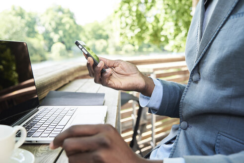 Geschäftsmann überprüft sein Telefon, während er auf einer Terrasse an seinem Laptop arbeitet, Teilansicht - IGGF00094