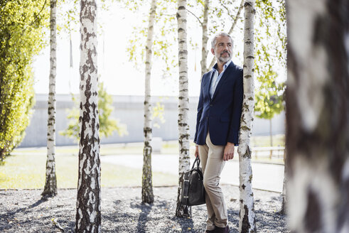 Businessman holding bag leaning against a tree - DIGF02654