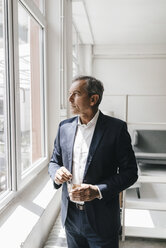Mature businessman with glass of coffee in his office looking out of window - KNSF02358