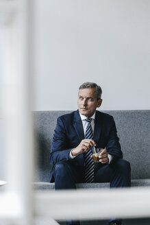 Älterer Geschäftsmann mit einem Glas Kaffee auf der Couch in seinem Büro sitzend und aus dem Fenster schauend - KNSF02383