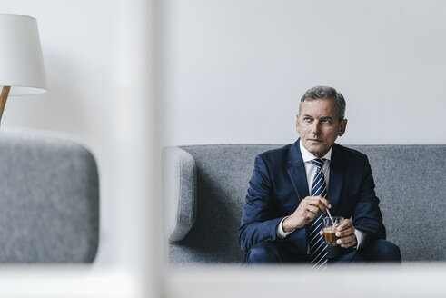 Älterer Geschäftsmann mit einem Glas Kaffee auf der Couch in seinem Büro sitzend - KNSF02384