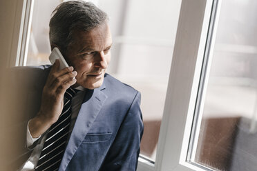 Lächelnder reifer Geschäftsmann, der mit seinem Handy aus dem Fenster schaut - KNSF02386