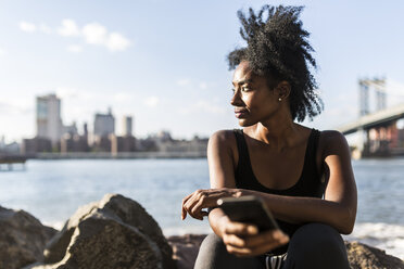 USA, New York City, Brooklyn, Frau mit Mobiltelefon am Wasser sitzend - GIOF03099