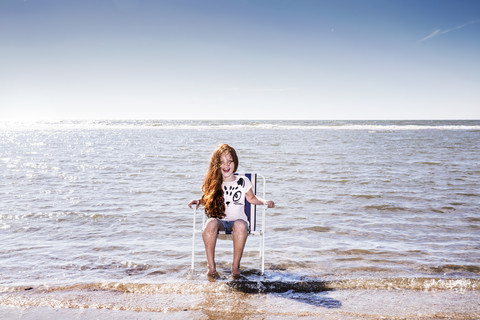 Niederlande, Zandvoort, glückliches Mädchen sitzt auf einem Stuhl im Meer, lizenzfreies Stockfoto