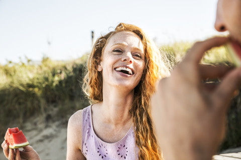 Niederlande, Zandvoort, glückliche Frau isst Wassermelone und schaut auf einen Mann am Strand, lizenzfreies Stockfoto