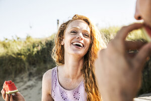 Niederlande, Zandvoort, glückliche Frau isst Wassermelone und schaut auf einen Mann am Strand - FMKF04319