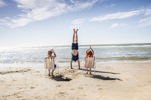 Netherlands, Zandvoort, children clapping hands for father doing a handstand on the beach - FMKF04374