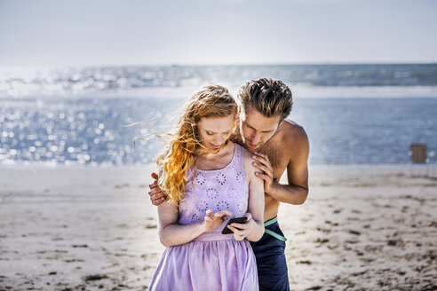 Niederlande, Zandvoort, Paar am Strand schaut auf Handy - FMKF04378