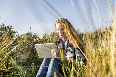 Netherlands, Zandvoort, smiling woman sitting in dunes with tablet - FMKF04382