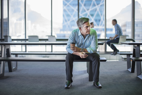 Geschäftsmann in einem modernen Büro auf einer Bank sitzend mit Kaffee zum Mitnehmen und Smartphone, lizenzfreies Stockfoto