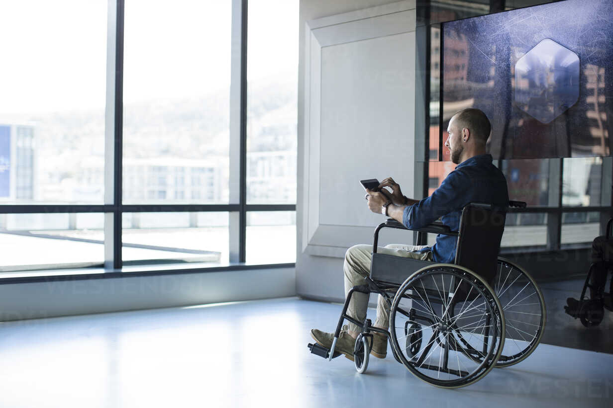 Mann im Rollstuhl, der im Büro aus dem Fenster schaut und ein Tablett  benutzt, lizenzfreies Stockfoto