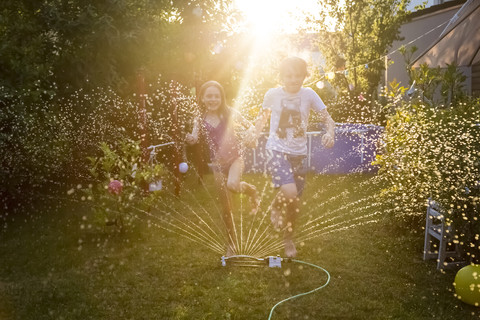 Brother and sister having fun with lawn sprinkler in the garden stock photo