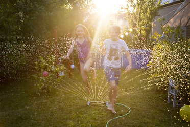 Brother and sister having fun with lawn sprinkler in the garden - SARF03349