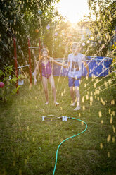 Brother and sister having fun with lawn sprinkler in the garden - SARF03350