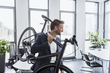 Smiling businessman with bicycle on his shoulder in the office - DIGF02678