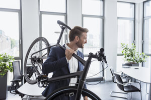 Lächelnder Geschäftsmann mit Fahrrad auf der Schulter im Büro - DIGF02678