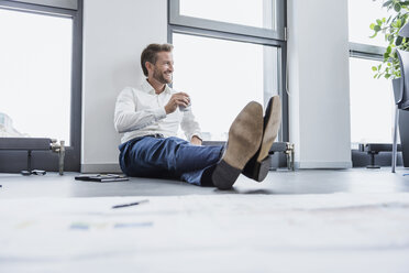 Entspannter Geschäftsmann, der mit einer Tasse Kaffee auf dem Boden seines Büros sitzt - DIGF02685