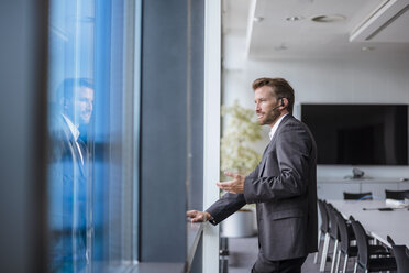 Relaxed businessman with bluetooth headset standing in his office looking out of the window - DIGF02704