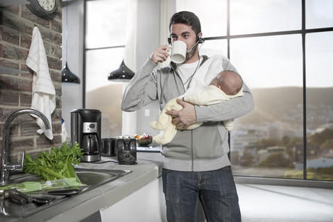Father with headset drinking coffee in kitchen holding baby stock photo