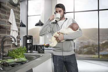 Vater mit Headset trinkt Kaffee in der Küche und hält sein Baby - ZEF14480
